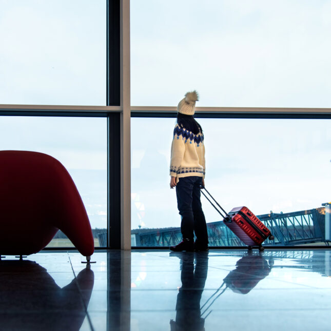 A person wearing a sweater and a beanie looks out the window at the airport with a red suitcase — first opinion coverage from STAT