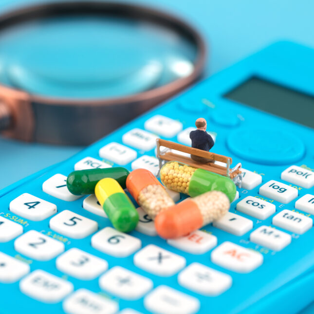 In front of five capsules, a miniature sits on a bench atop a calculator next to a magnifying glass — first opinion coverage from STAT