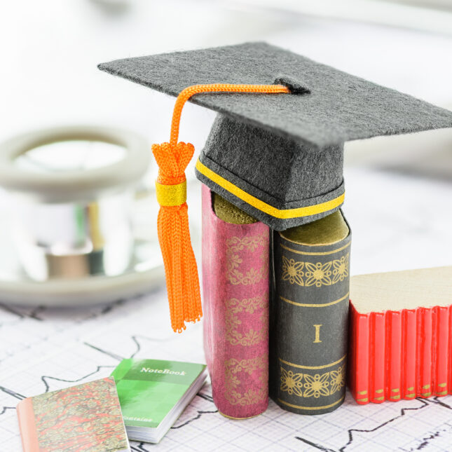 A graduation cap, placed atop miniature books, rests on a piece of paper imprinted with a heartbeat diagram. A stethoscope lies behind the books — first opinion coverage from STAT