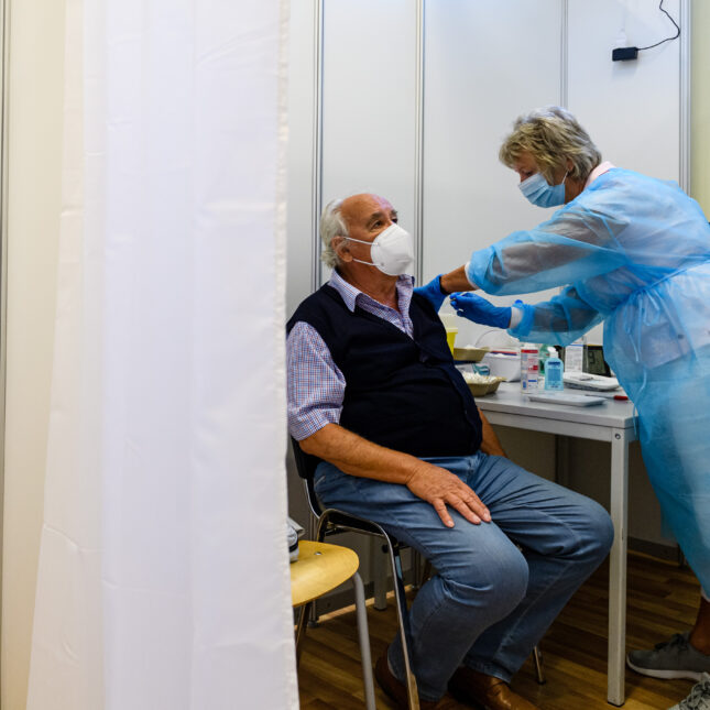 Behind a curtain, a medical staff wearing PPE administrates a COVID-19 booster shot to a senior who is masked and sitting down — health coverage from STAT