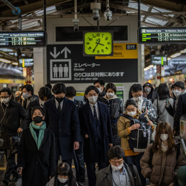 Dozens of commuters, wearing face masks, swarm toward a descending escalator at a train station in Tokyo, Japan — in the lab coverage from STAT
