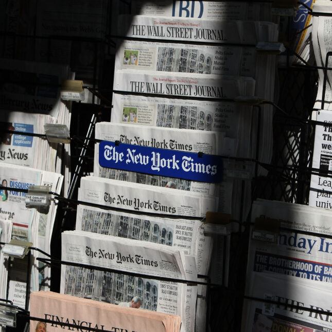 The sun shines on a newsstand displaying newspapers from The New York Times, The Wall Street Journal, Financial Times — coverage from STAT